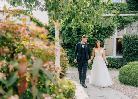 A couple holding hands, walking in a lush garden of a charming hotel.