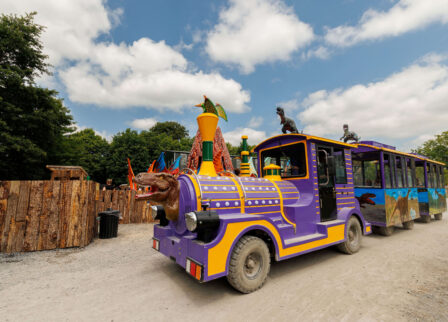 A colourful train with dinosaur decorations rides through a scenic park under a blue sky.