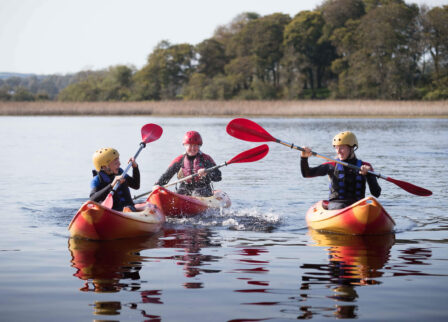 A family enjoying kayaking together on a serene lake with lush trees in the background.