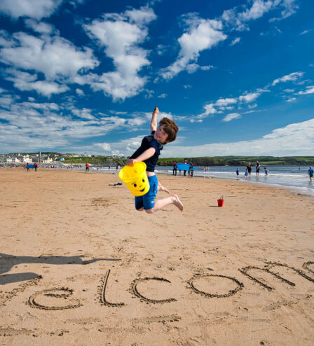 Child jumping on beach with "Welcome" in sand, blue sky and sea in background.