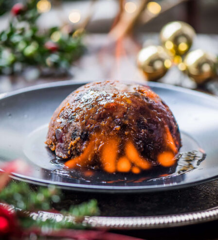 A flaming Christmas pudding on a festive table with holly and bauble decorations.