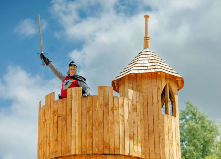 Child dressed as a knight brandishing a sword on a wooden castle tower under a blue sky.