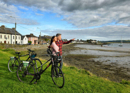 A couple with bicycles enjoying a scenic coastal view, pointing towards boats on the sea.
