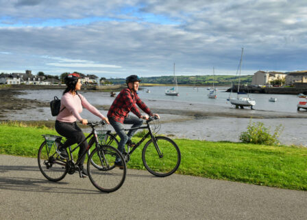 A couple cycling by a scenic harbour with moored boats and green landscape.