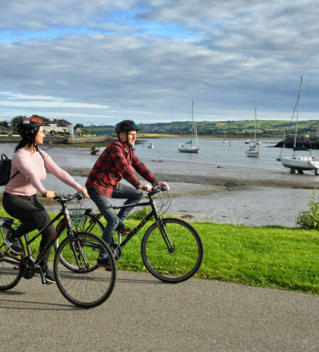 A couple cycling by a scenic harbour with moored boats and green landscape.