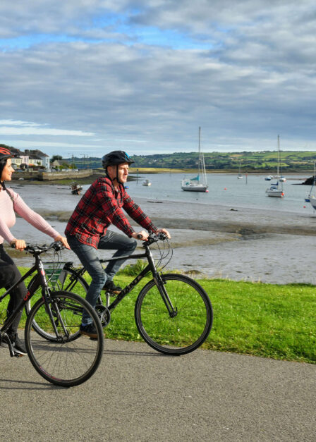 A couple cycling by a scenic harbour with moored boats and green landscape.