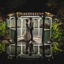 A couple embraces under a lit gazebo at night beside a reflective pond in a garden setting.