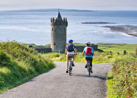 Couple cycling towards a historic coastal tower with a scenic ocean view.