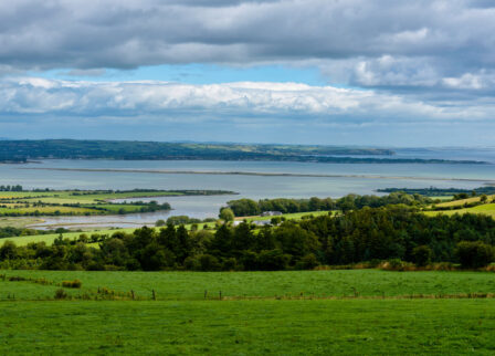 A peaceful countryside landscape with lush green fields and a distant view of the sea under a cloudy sky.