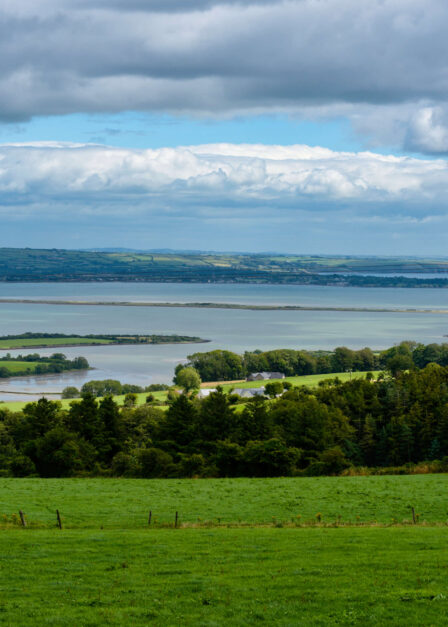 A peaceful countryside landscape with lush green fields and a distant view of the sea under a cloudy sky.