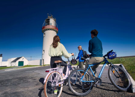 Family cycling near a scenic lighthouse under a blue sky.