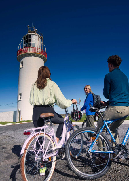 Family cycling near a scenic lighthouse under a blue sky.