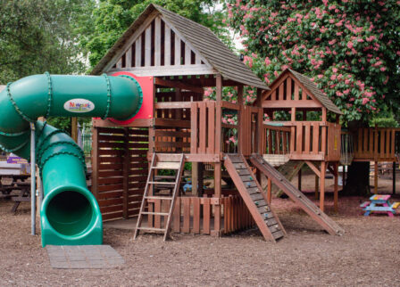 Families enjoying outdoor wooden playground at a vibrant hotel or resort garden.
