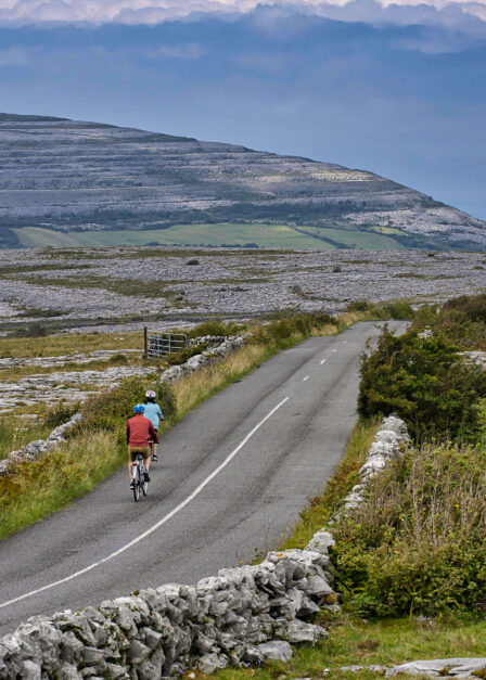 Couple cycling along scenic countryside road with rolling hills and distant sea views.