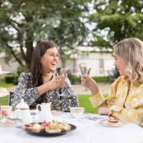 Two women enjoying afternoon tea in a garden, raising glasses in a toast.