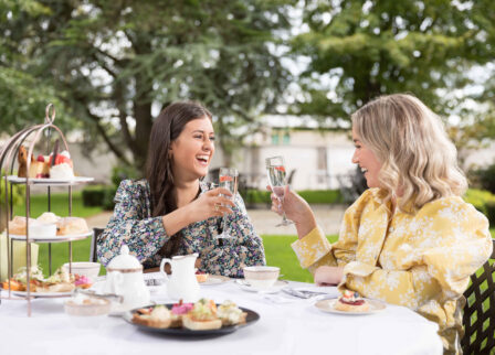 Two women enjoying afternoon tea in a garden, raising glasses in a toast.