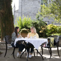 A couple enjoying a sunny outdoor brunch at a hotel garden with lush greenery and stone architecture.