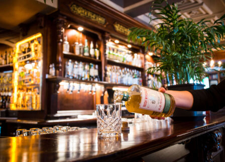 A cosy bar with a person pouring whisky into a glass, surrounded by warm lighting.