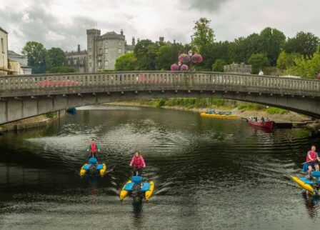 Families enjoying hydrobikes on a scenic river under a historic stone bridge near a castle.