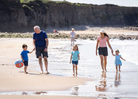 Family enjoying a sunny beach walk, children with ball and waves gently lapping.