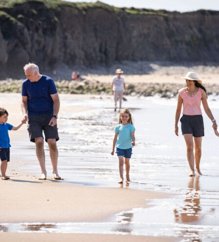 Family enjoying a sunny beach walk, children with ball and waves gently lapping.