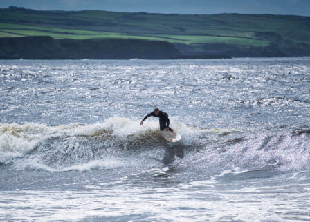 A surfer rides a wave at a scenic coastal location, under a cloudy sky.
