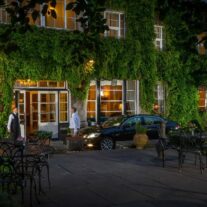A couple arrives at a charming, ivy-covered hotel entrance at dusk, greeted warmly by the staff.