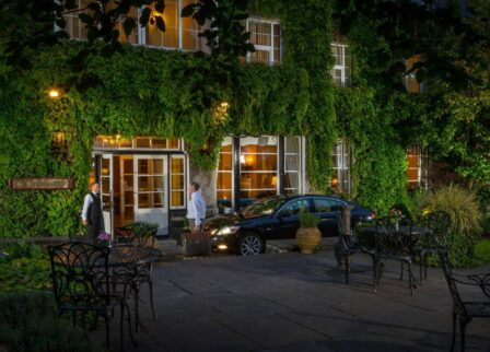 A couple arrives at a charming, ivy-covered hotel entrance at dusk, greeted warmly by the staff.